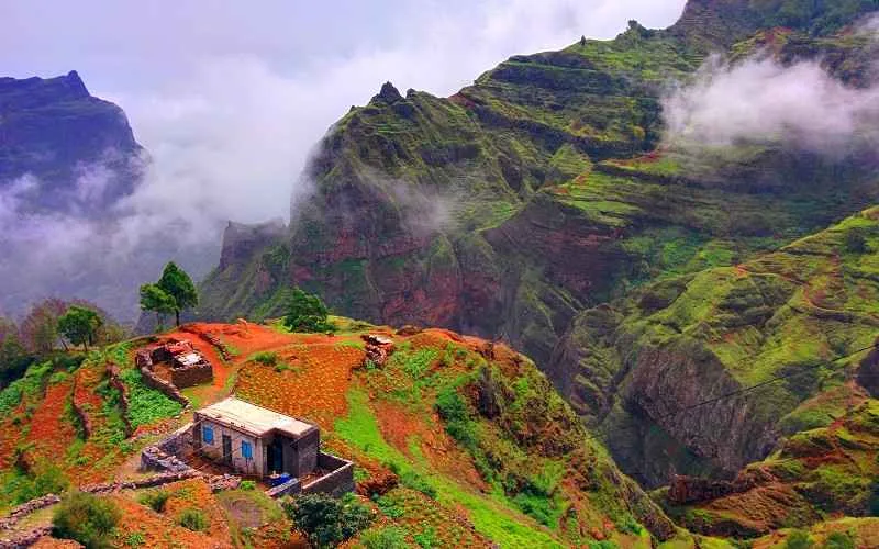 Une maison isolée en haut d'une faaise, entourée de montagnes immenses et de verdure